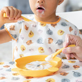 A young child in a colorful bib eats from a plate with Bumkins Silicone Dipping Spoons: Winnie the Pooh, while an adult assists.