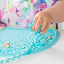 Load image into Gallery viewer, Close-up of a child's hand holding a Bumkins Silicone Chewtensils®: Blue on a blue silicone placemat with cereal pieces.
