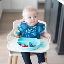 Load image into Gallery viewer, A curly-haired baby in high chair, wearing a blue bib, uses Bumkins Silicone Chewtensils®: Blue to eat fruit from a blue divided plate.
