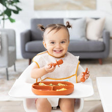 Load image into Gallery viewer, A toddler with pigtails in a high chair wearing a beige bib uses Bumkins Silicone Chewtensils®: Clay to eat from a divided plate.

