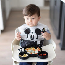 Load image into Gallery viewer, A toddler wearing a black &amp; white bib in a high chair uses Bumkins Silicone Chewtensils®: Mickey Mouse to eat from a Mickey-shaped plate.
