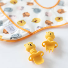 Load image into Gallery viewer, Close-up of Bumkins Silicone Chewtensils®: Winnie The Pooh on a white background, with a colorful printed bib blurred behind.
