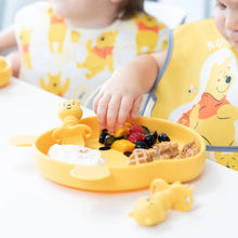 Load image into Gallery viewer, Close-up of a child in a bear bib eating fruit from a yellow plate with Bumkins Silicone Chewtensils®: Winnie The Pooh nearby.

