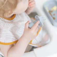 Load image into Gallery viewer, Close-up of a toddler in a high chair wearing a beige bib and holding a Bumkins Silicone Chewtensils®: Gray over a gray plate.
