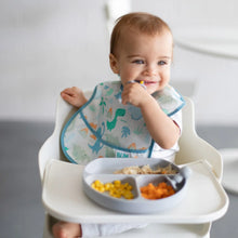 Load image into Gallery viewer, A baby wearing a dinosaur bib in a high chair uses Bumkins Silicone Chewtensils®: Gray to explore baby-led weaning from a gray plate.
