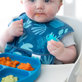Close-up of a baby in a blue bib enjoying food from a blue plate with Bumkins Silicone Chewtensils®: Blue Jelly.