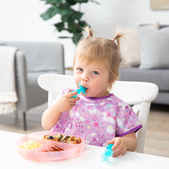 In a cozy living room, a toddler in a purple unicorn bib uses Bumkins Silicone Chewtensils®: Blue Jelly for baby-led weaning.