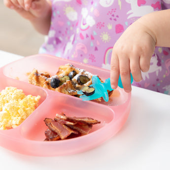 A child enjoys waffles with blueberries using Bumkins Silicone Chewtensils®: Blue Jelly from a pink plate with eggs and bacon.