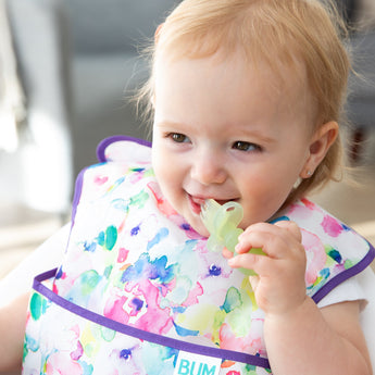 A girl in a floral bib places a Bumkins Silicone Chewtensils®: Green Jelly in her mouth while smiling.