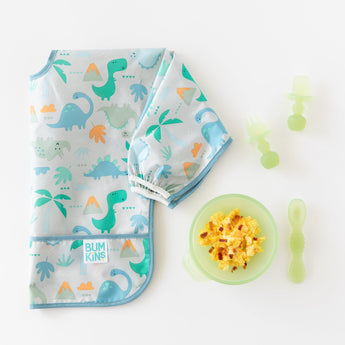 A folded dinosaur-print bib lays next to a green bowl with eggs and Bumkins Silicone Chewtensils®: Green Jelly on a white background.