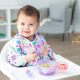 A baby in a high chair smiles, wearing a floral bib and holding Bumkins Silicone Chewtensils®: Purple Jelly by a purple bowl.