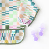 Bumkins Silicone Chewtensils®: Purple Jelly rest near a colorful sleeved bib on a white background.