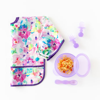 Bumkins Silicone Chewtensils®: Purple Jelly sit next to a purple bowl & spoon with spaghetti and a folded floral bib on a white background.