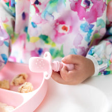 Load image into Gallery viewer, Close-up of a child in a colorful bib holding a Bumkins Silicone Chewtensils®: Pink next to a pink divided plate.
