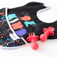 Load image into Gallery viewer, A colorful baby bib with abstract designs is paired with Bumkins Silicone Chewtensils®: Red on a white background.
