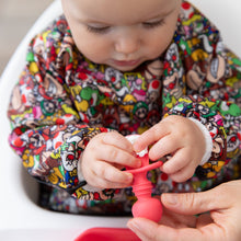 Load image into Gallery viewer, A baby wearing a Super Mario™ Mashup sleeved bib, touches a Bumkins Silicone Chewtensils®: Red with cottage cheese on it.
