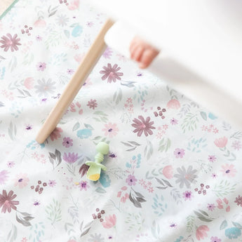 A Bumkins Silicone Chewtensils®: Sage with applesauce lays on a floral mat underneath a high chair with a baby's foot visible.