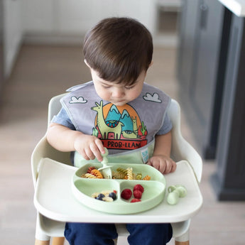 A toddler wearing a llama bib in a high chair uses Bumkins Silicone Chewtensils®: Sage to enjoy pasta and berries from a green plate.