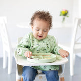 A toddler sits in a high chair, wearing a green bib and using Bumkins Silicone Chewtensils®: Sage to eat from a green divided plate.