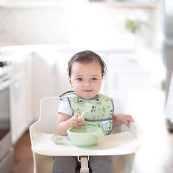 A baby grins in a high chair, wearing a green bib and holding Bumkins Silicone Chewtensils®: Sage near a matching bowl.