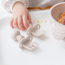 Load image into Gallery viewer, Close-up of a child's hand grabbing Bumkins Silicone Chewtensils®: Sand on a high chair tray with a bowl of food nearby.
