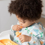 A curly-haired toddler in a high chair wearing a dinosaur bib enjoys a meal using Bumkins Silicone Chewtensils®: Tangerine.