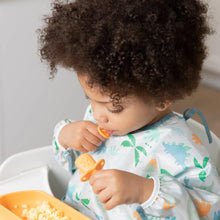 Load image into Gallery viewer, A curly-haired toddler in a high chair wearing a dinosaur bib enjoys a meal using Bumkins Silicone Chewtensils®: Tangerine.
