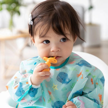 A toddler in a blue sea animal-themed bib chews a Bumkins Silicone Chewtensils®: Tangerine.