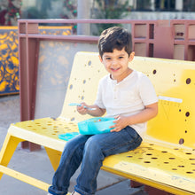 Load image into Gallery viewer, A young boy sits on a yellow bench, holding Bumkins Spoon + Fork: Blue and a blue lunch box.
