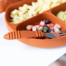 Load image into Gallery viewer, Close-up of an orange divided plate with fruit, pasta, avocado, with a Bumkins Spoon + Fork: Clay resting on the edge.

