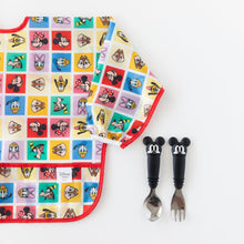 Load image into Gallery viewer, A Bumkins Sleeved Bib: Mickey &amp; Friends Check Mates lays flat on a white background next to Spoon + Fork: Mickey Mouse.
