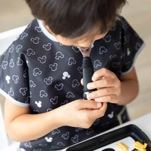 Load image into Gallery viewer, A child in a Mickey Mouse bib holds a Bumkins Spoon + Fork: Mickey Mouse in their mouth at a table with a black plate nearby.
