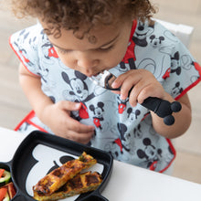 Load image into Gallery viewer, A boy in a Mickey Mouse bib uses Bumkins Spoon + Fork: Mickey Mouse to eat a meal with a Mickey Mouse Grip Dish at a white table. 
