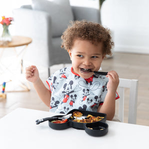 A boy in a Junior Bib: Mickey Mouse Classic sits at a white table holding Mickey Mouse fork in his mouth with the Mickey Grip Dish on table.