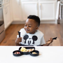 Load image into Gallery viewer, A child in a Mickey Mouse bib holds Bumkins Spoon + Fork: Mickey Mouse at a white table with a Mickey Mouse-shaped plate of food.
