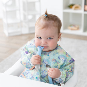 A baby with a topknot smiles in a high chair, holding Bumkins Spoon + Fork: Stitch in a bright playroom.