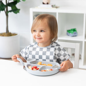 A toddler in a checkered bib smiles, using Bumkins Spoon + Fork: Gray to eat from a gray divided plate at a white table.