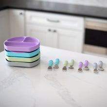 Load image into Gallery viewer, Bumkins Spoon + Fork: Lavender sits among other utensil sets and a stack of silicone divided plates on a marble kitchen counter.
