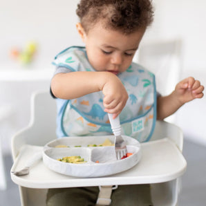A toddler in a high chair wearing a dinosaur bib uses Bumkins Spoon + Fork: Marble to eat from a matching plate.