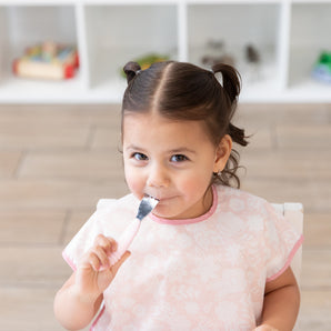 Child wearing a Bumkins Junior Bib: Floral, eating from a Bumkins Spoon + Fork: Pink.