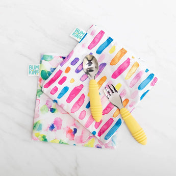 Bumkins Spoon + Fork: Pineapple lays on top of two colorful waterproof pouches on a marble background.