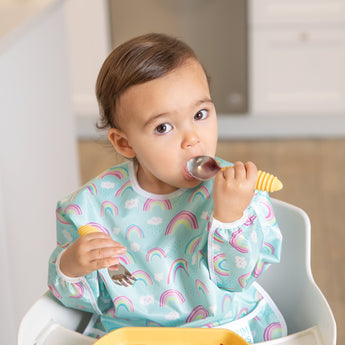 A toddler in a rainbow-print sleeved bib sits in a high chair, holding Bumkins Spoon + Fork: Pineapple while looking up at the camera.