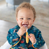 A smiling baby wearing a blue animal-patterned bib chews a Bumkins Spoon + Fork: Pineapple while seated in a high chair.