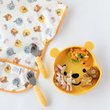 Bumkins Spoon + Fork: Pineapple lays on top of a colorful bib next to a bear-shaped plate of food on a white background.