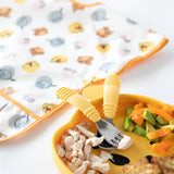 Close-up of Bumkins Spoon + Fork: Pineapple on a yellow plate with chicken, avocado & veggies near a colorful bib on a white background.