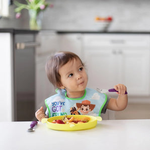 A toddler in a bib uses Bumkins Spoon + Fork: Purple to eat fruit and pasta from a yellow plate at a white kitchen table.