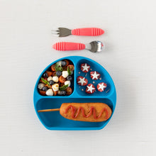 Load image into Gallery viewer, A dark blue divided plate with a corn dog, salad, and fruit sits underneath Bumkins Spoon + Fork: Red on a white background.
