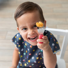 Load image into Gallery viewer, A smily boy in a Bumkins Junior Bib: Super Mario™ Lineup sits happily in a white chair holding Red Spoon + Fork Set in front of his face.
