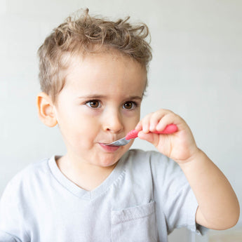 A curly-haired boy with a curious expression holds a Bumkins Spoon + Fork: Red in his mouth.