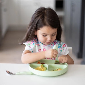 A young girl with long brown hair in a floral bib uses Bumkins Spoon + Fork: Sage to eat from a matching plate at a white table.